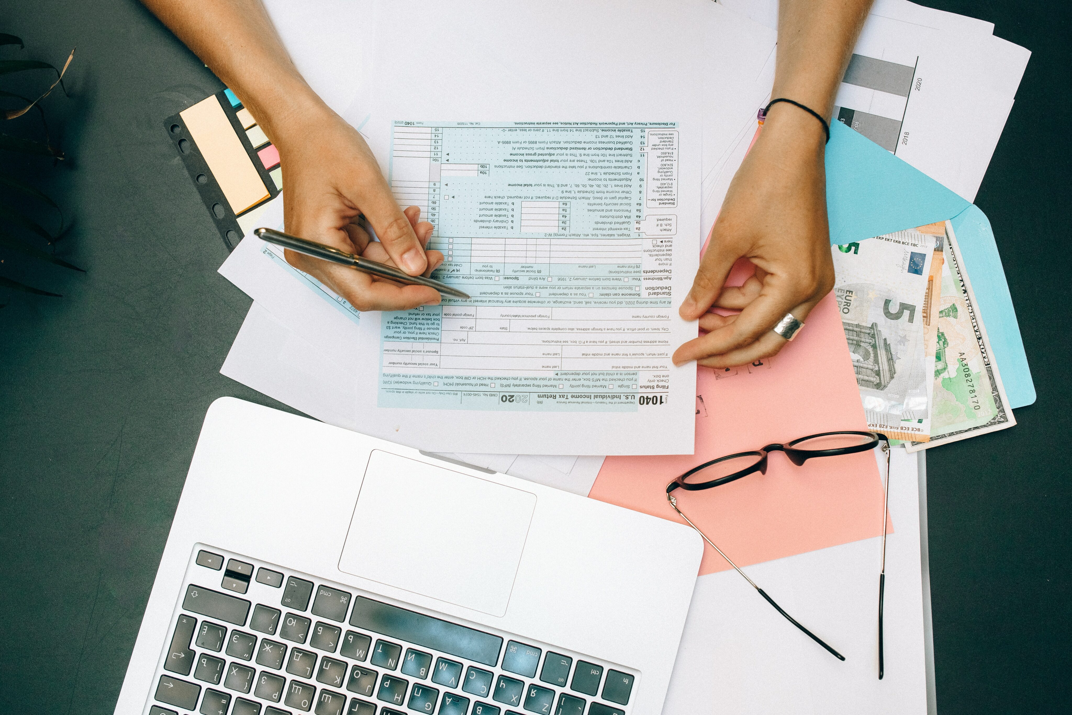 Person's hands with pen and laptop, glasses, tax forms, money and papers on desk.