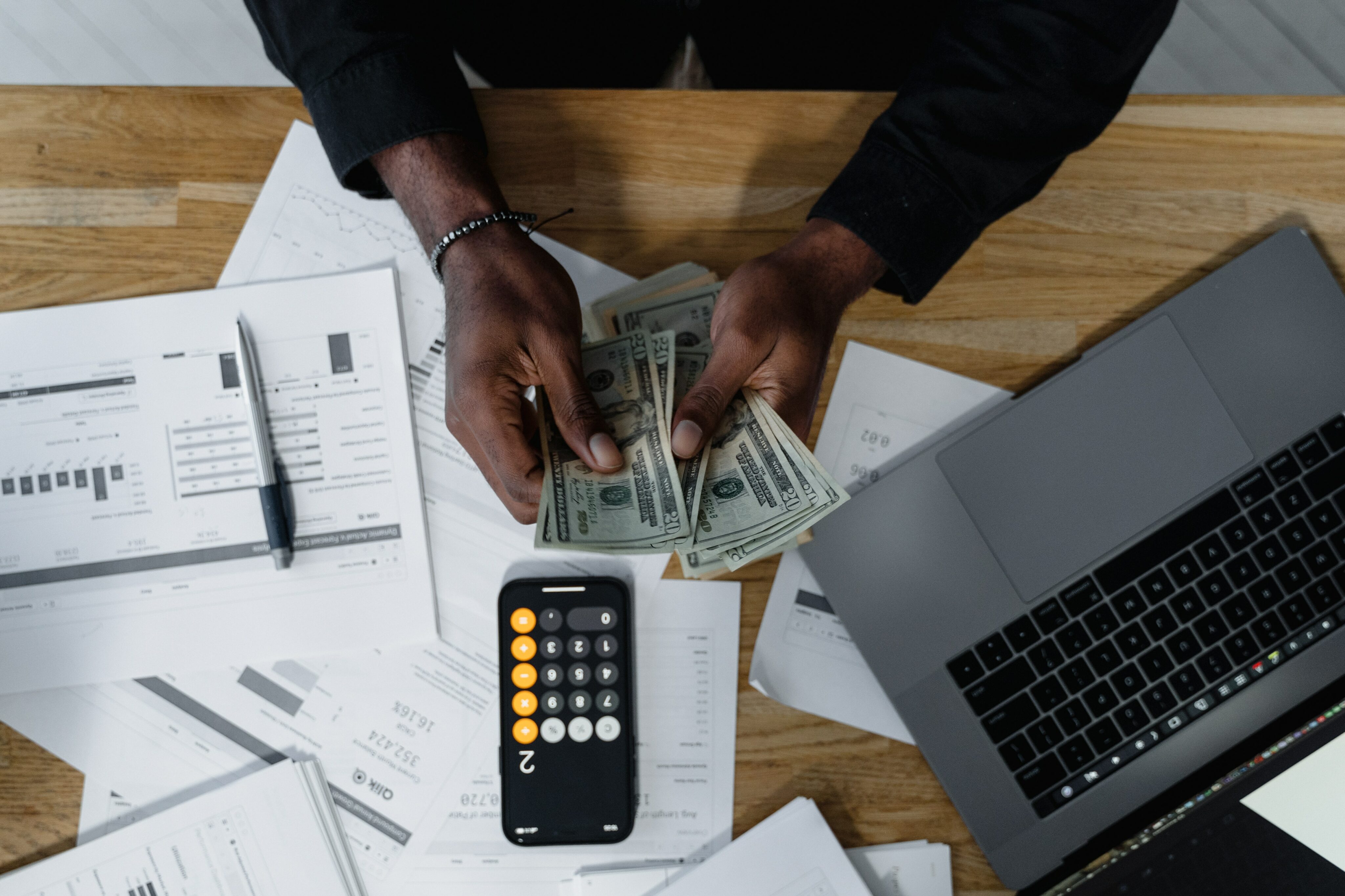 Person's hands holding money with calculator, laptop, pen, and various papers with charts, graphs, and calculations on table