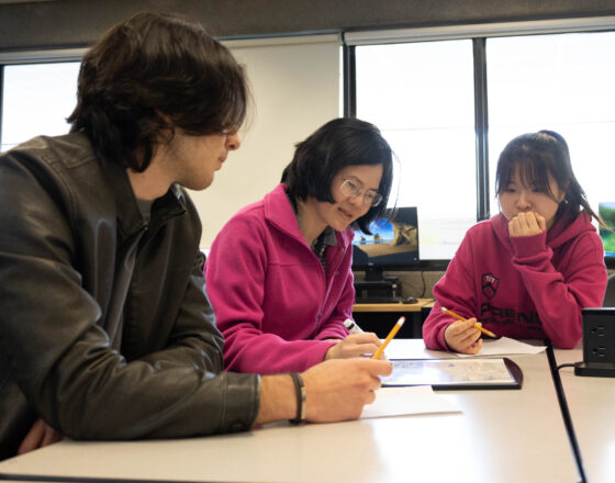 Two students take notes while a tutor writes on a tabletop whiteboard.