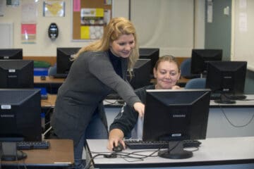 student sitting a desk with a desktop while teacher stands next to them and points at the screen