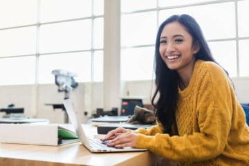 Women sitting in a desk smiling at the camera while working on a laptop in an office
