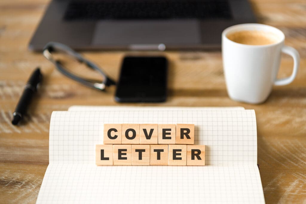 A close up photo of a desk with a laptop, glasses, phone, pen, mug, and lined paper with wooden blocks that spell out cover letter