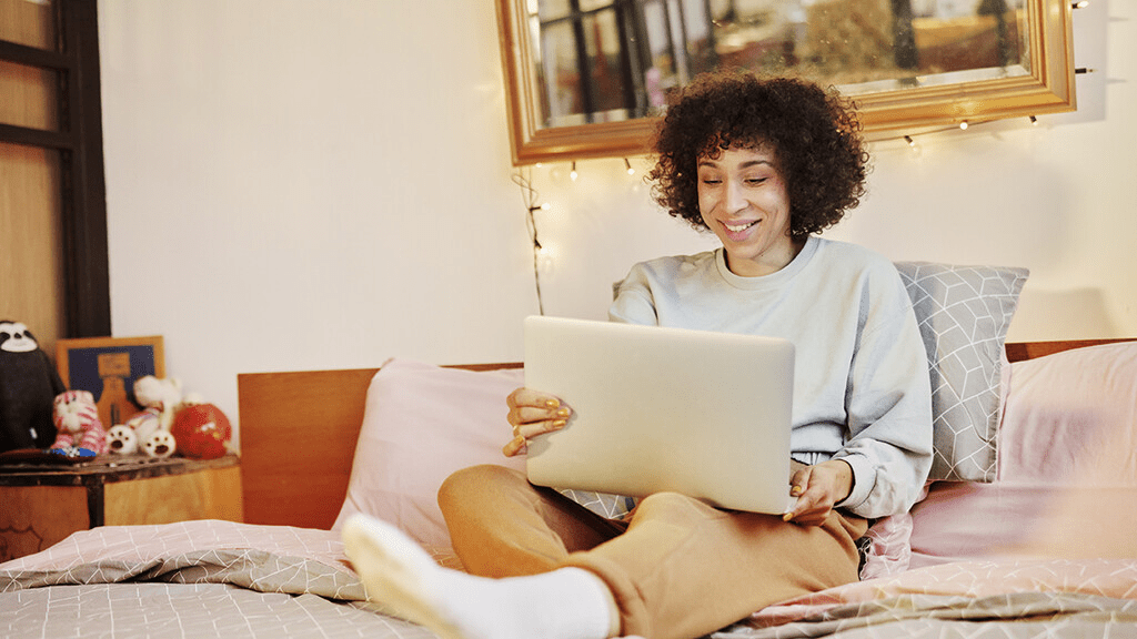 A women smiling at her laptop while sitting in bed