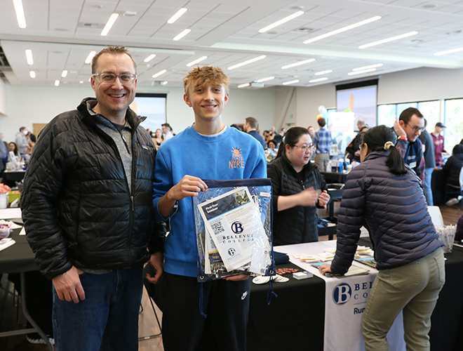 A father and son stand next to one another and smile while looking at the camera. In the background, people walk around a large room filled with tables.