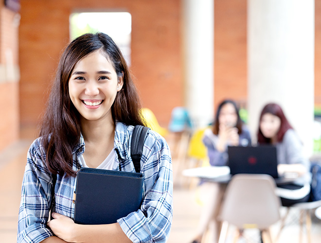A young woman looks at the camera smiling. She is wearing a backpack and carrying a notebook. In the background, students sit at a table looking at a laptop.
