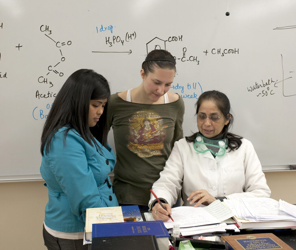 two female-presenting students observe a seated female-presenting person wearing a white lab coat and goggles, holding a pen