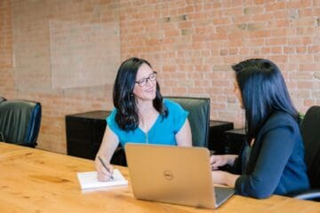 Two women sitting at a desk and talking with a laptop and one woman is taking notes