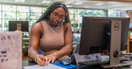 A student makes notes on paper while working in front of a tablet and desktop computer.