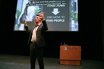 A speaker gestures in front of a projection screen and podium while delivering a presentation.