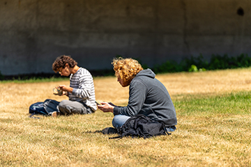 Students sit cross-legged on the grass and look at their phones.