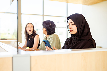 Two students discuss in a common area while a third uses their phone.