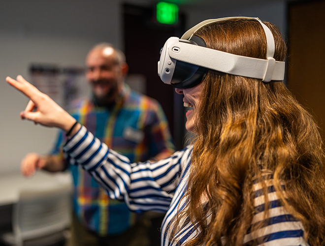 A student laughs in delight while wearing a VR headset and gesturing with their hand.
