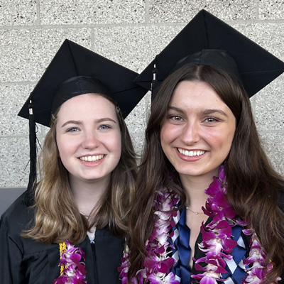 Two female students wearing graduation caps and pink flower leis smile for the camera