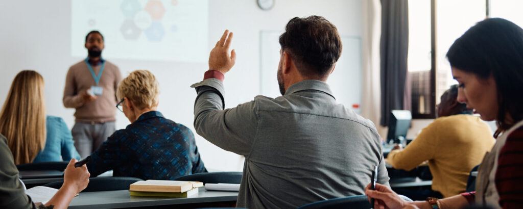 A man raises his hands in class while other students take notes and teacher is in the front addressing the class.