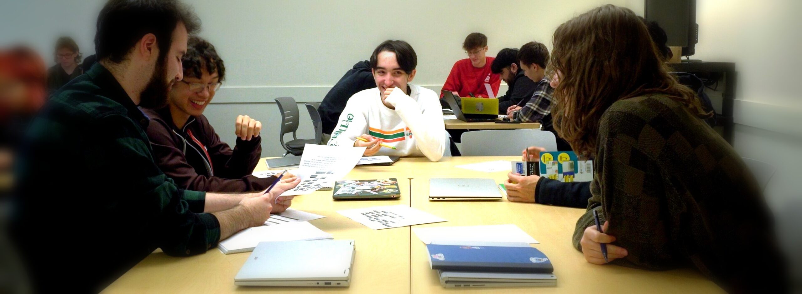 Students seated around tables collaborate in a classroom, reviewing printed materials and laptops during a group discussion.