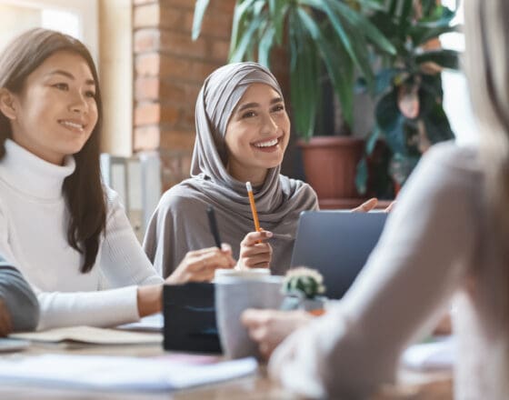 focus is on two women who are smiling in what seems to be a meeting. One is wearing a turtle neck and the other has a hijab