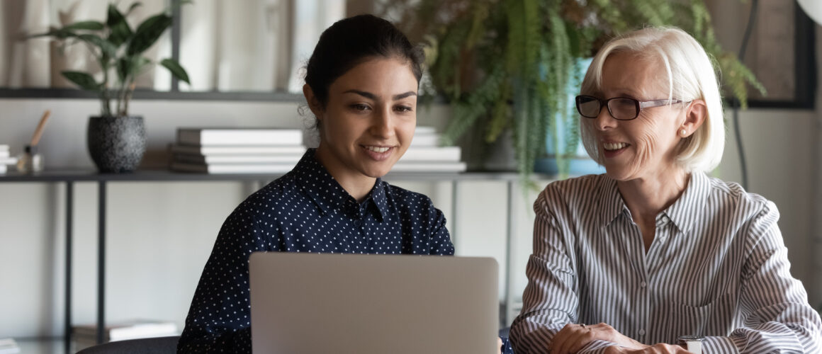 A younger intern and an older colleague sit together at a desk, smiling while reviewing information on a laptop in a modern office.