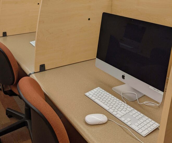 Apple desktop computer in library carrel with red rolling chair. The carrel is in a row with other carrels.