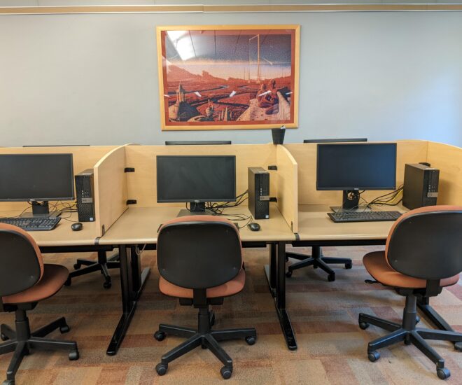 3 computers on carrels in the library. Red rolling chairs sit in front of them. A piece of art hangs on the wall in the background.