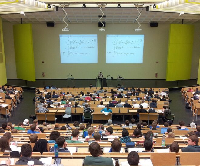 Large auditorium college classroom from back of class, shows students looking at two projected screens.