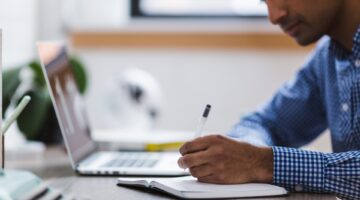 Young man writing on a notepad. He is sitting next to an open laptop.