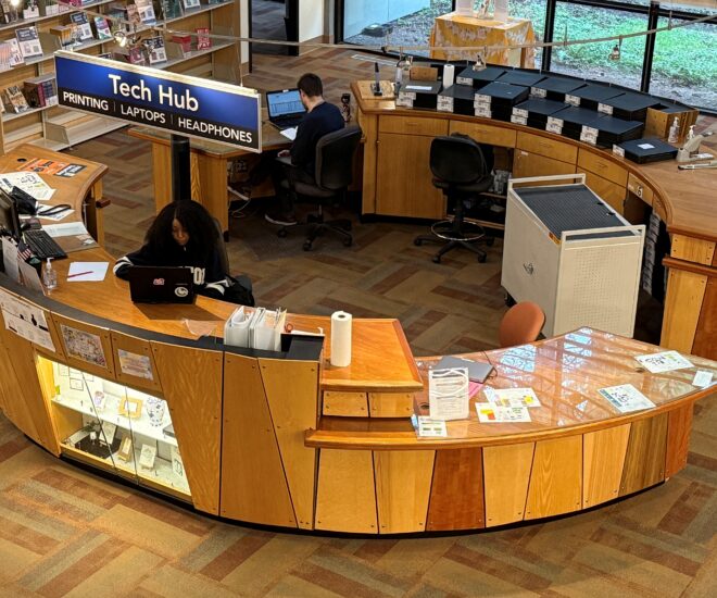 2 Students sit looking at laptops at the Tech Hub desk in the library. A Sign overhead reads Tech Hub printing, laptops, headphones.