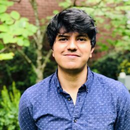 Person wearing a blue patterned shirt standing outdoors in front of green foliage and a brick wall.