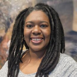 Person with long, dark locs wearing a light gray textured top, standing in front of a softly blurred, earthy-toned background.