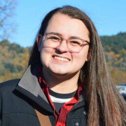 Person outdoors wearing a black jacket and red plaid shirt, with a forested hillside and blue sky in the background.