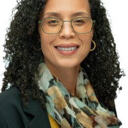 Person with curly hair wearing a patterned scarf, yellow top, and dark blazer against a plain background.