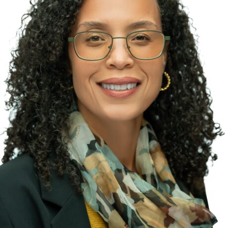 Person with curly hair wearing a patterned scarf, yellow top, and dark blazer against a plain background.