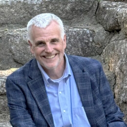 A man smiles in a headshot wearing a blazer in front of a stone wall.
