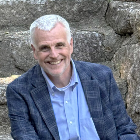 A man smiles in a headshot wearing a blazer in front of a stone wall.