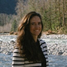 A woman smiles while standing outdoors near a river with trees in the background.