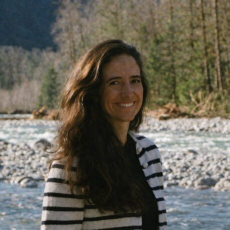 A woman smiles while standing outdoors near a river with trees in the background.