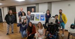 A group poses indoors around a board displaying XR project posters, with one participant using a powered wheelchair.