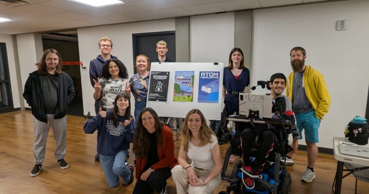 A group poses indoors around a board displaying XR project posters, with one participant using a powered wheelchair.