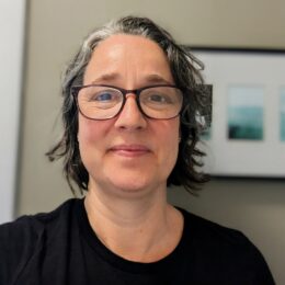 Close-up portrait of a person with short dark hair with gray streaks, wearing glasses and a black shirt, looking at the camera indoors