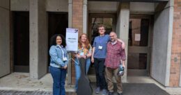 Four people stand outside Meany Studio Theatre next to a welcome sign, one holding a guitar case.