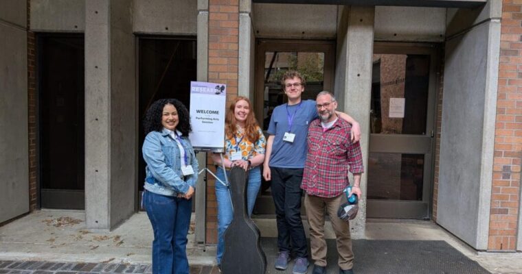 Four people stand outside Meany Studio Theatre next to a welcome sign, one holding a guitar case.