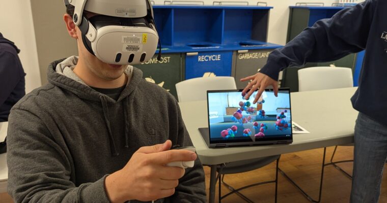 A person wearing a VR headset holds a controller while another gestures toward a laptop showing a 3D model.