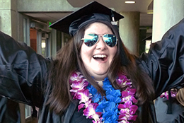 A recent BC grad celebrates while wearing sunglasses, cap and gown, and two leis.