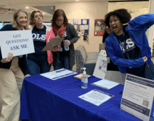 A member of the OLS community holds a sign that says "Got questions? Ask me" while others pose and gesture excitedly.
