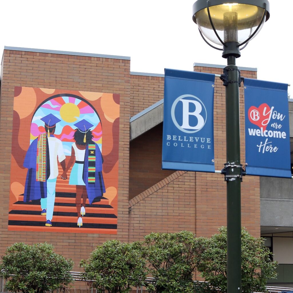 Image of wheatpaste mural Unity by Stat the Artist for Addressing Anti-Blackness Taskforce, shown beside a banner for Bellevue College. The mural depicts two of Black students in graduation regalia walking hand-in-hand up a flight of stairs toward a colorful horizon. 