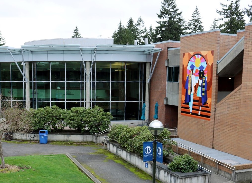 A wide view an outdoor courtyard of Bellevue College showing the mural Unity by Stat the Artist for Addressing Anti-Blackness Taskforce, and the building adjacent. 