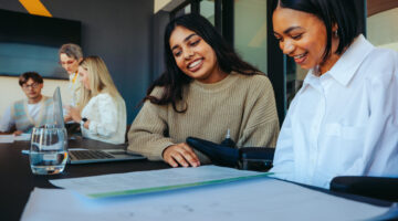 Two students looking at a paper and smiling with other students in the background grouped up togther.
