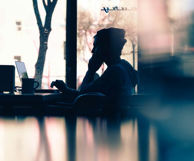 silhouette of a person in a beanie sitting in a cafe with their laptop and a mug