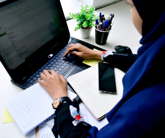 woman in headscarf at a desk on a laptop with notebooks, papers, and a phone