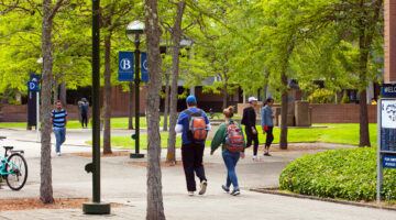 Students walking along a tree‑lined campus pathway with buildings, signs, and bicycles in the background.