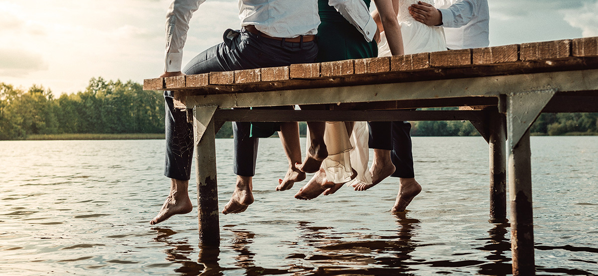 A group of friends sit on the edge of a dock, with their legs dangling toward the water.